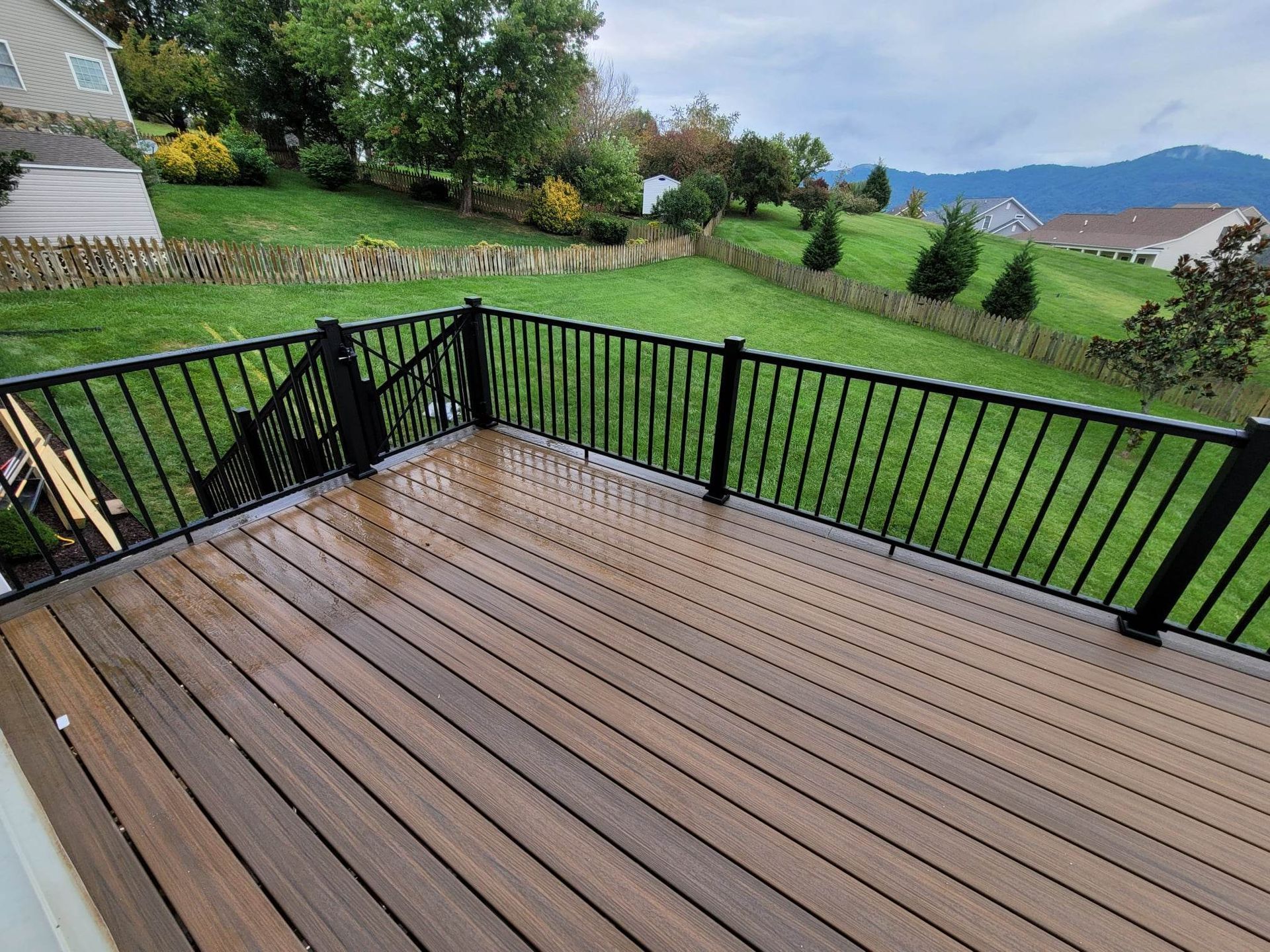 A high-angle view of a brown composite deck with a black metal railing overlooking a grassy backyard and distant hills.
