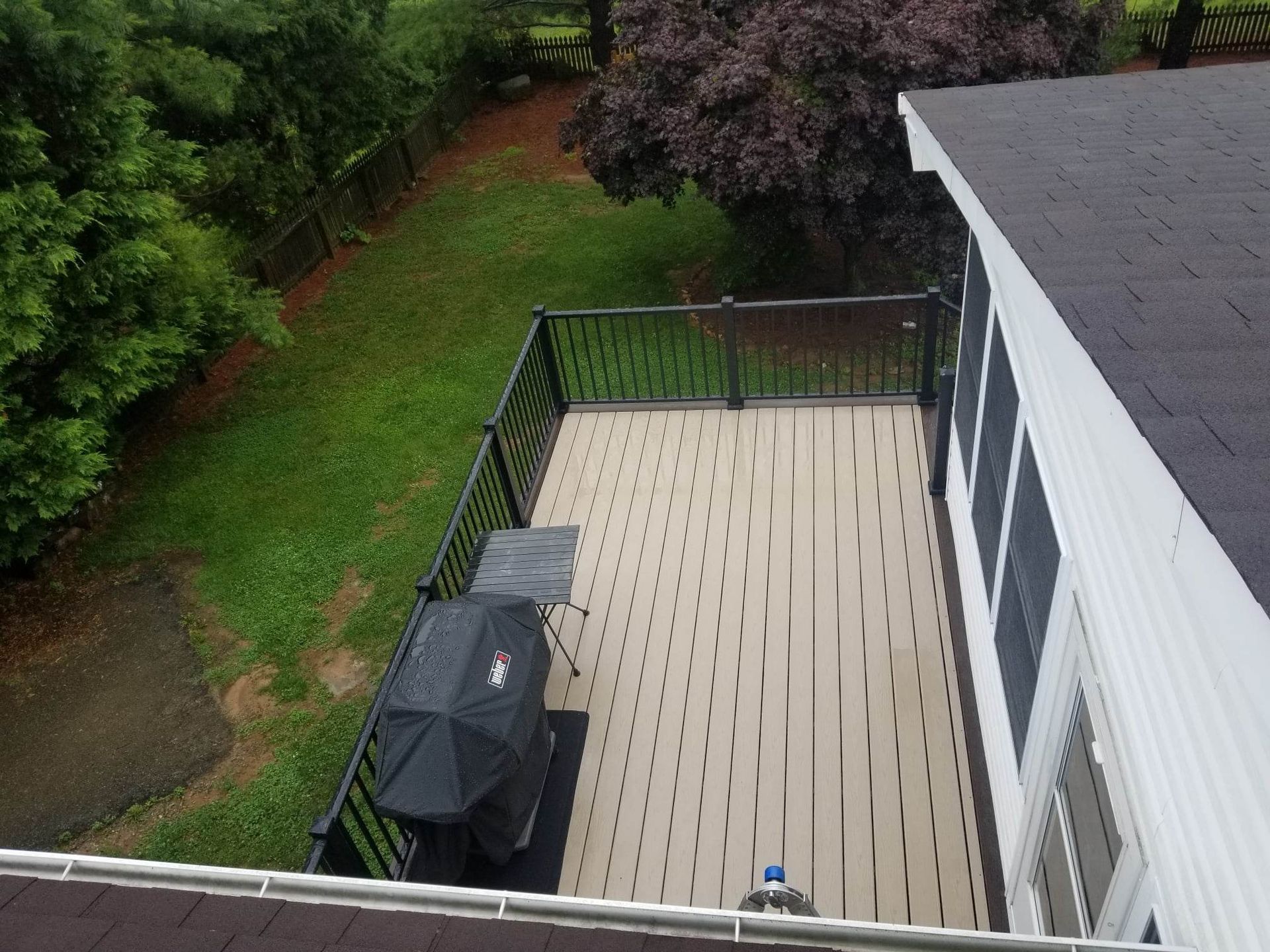 A high-angle view of a rectangular, light-colored deck with black railings next to a house, overlooking a green backyard.