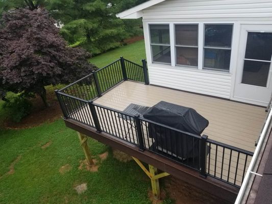 An elevated wooden deck with black railings and a covered grill, attached to a house next to a large purple-leaf tree.