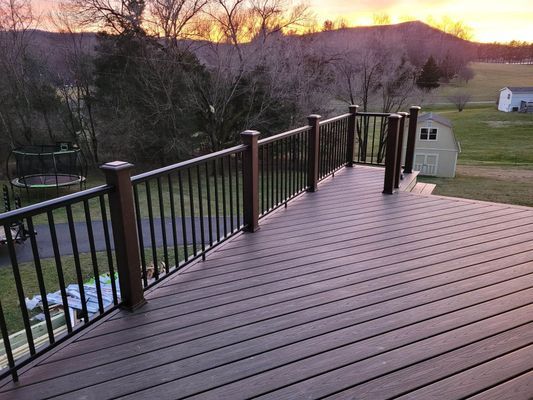 A brown composite deck with a black railing overlooks a grassy backyard at sunset.