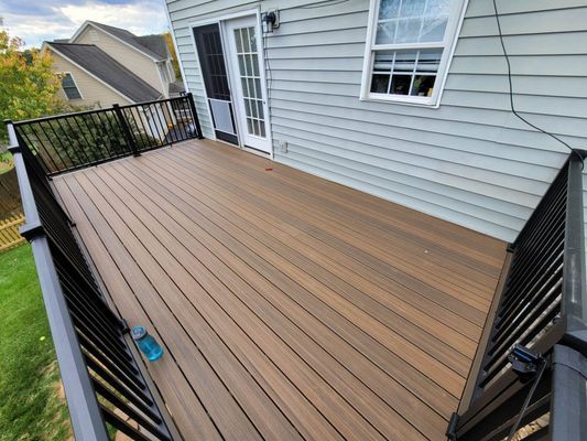 An elevated outdoor deck with brown composite decking boards and black railings, attached to the side of a grey house.