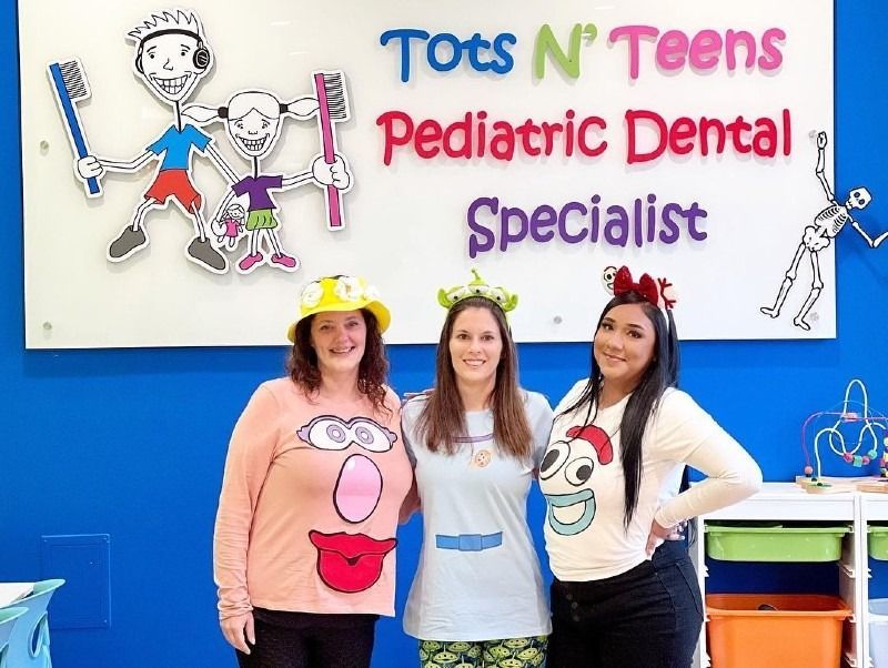 Three women are posing for a picture in front of a sign that says tots n teens pediatric dental specialist
