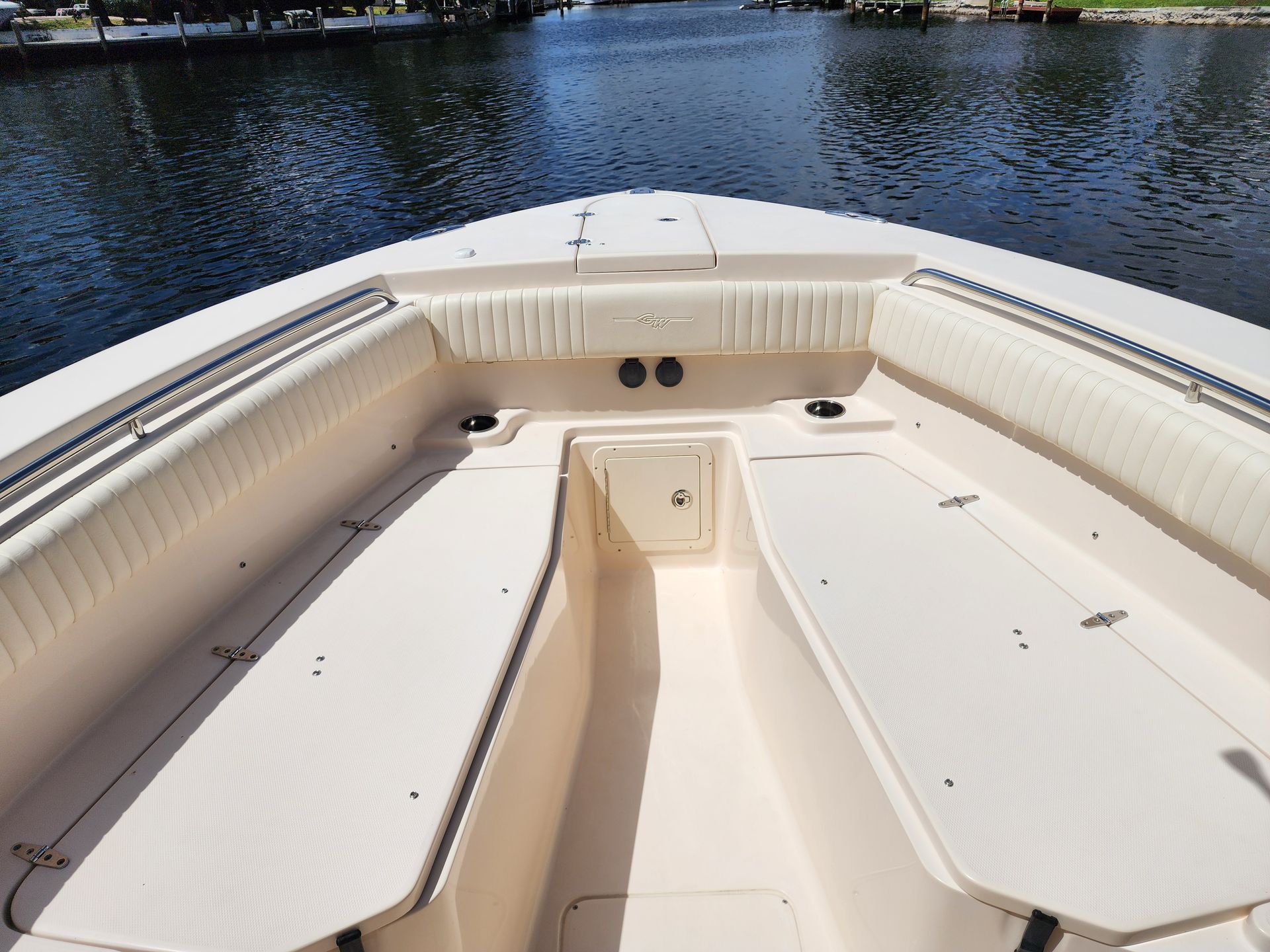 Bow of a boat with light-colored seating and storage, facing a body of water on a sunny day.