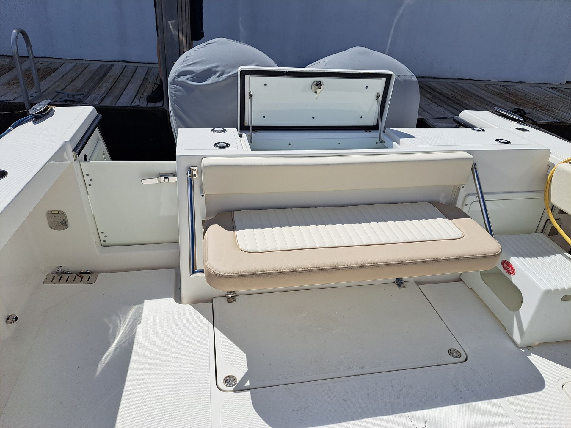 Boat's stern with a fold-down seat and storage compartment open. White deck and beige seat are visible.