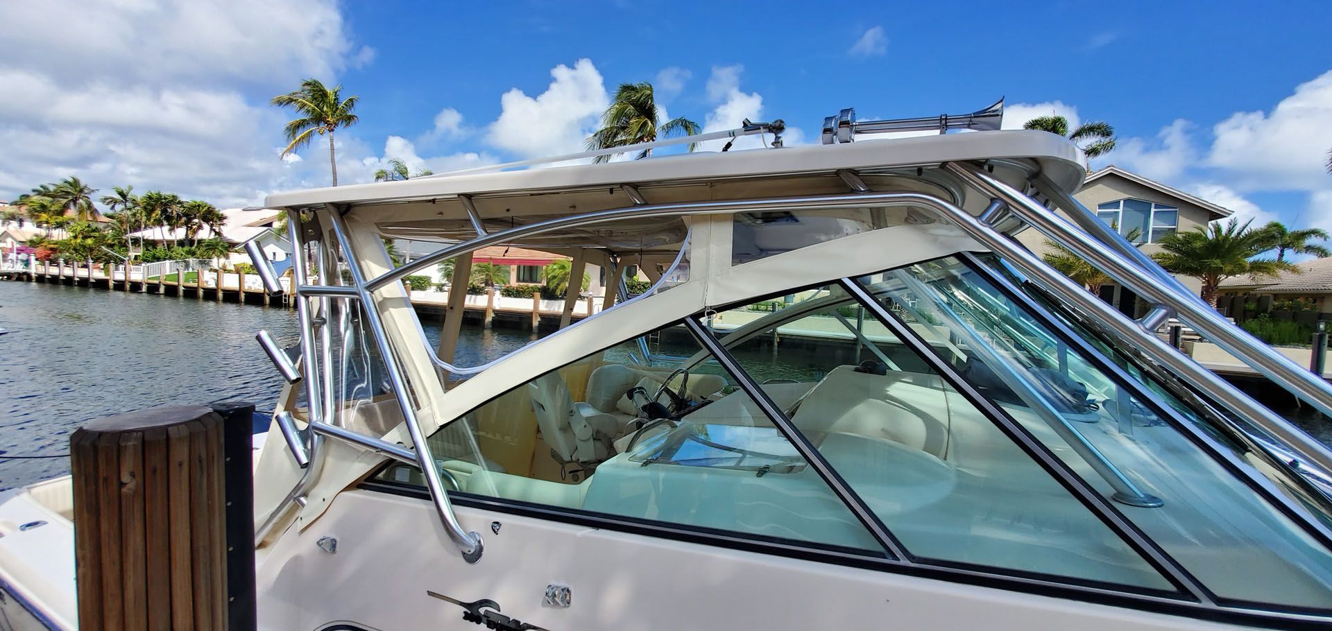 A boat with a windshield is docked near a wooden pier. Houses line the waterway under a blue sky.