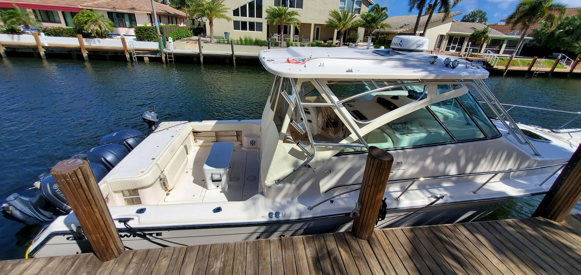 Boat docked at a pier, light blue water. Houses in background.