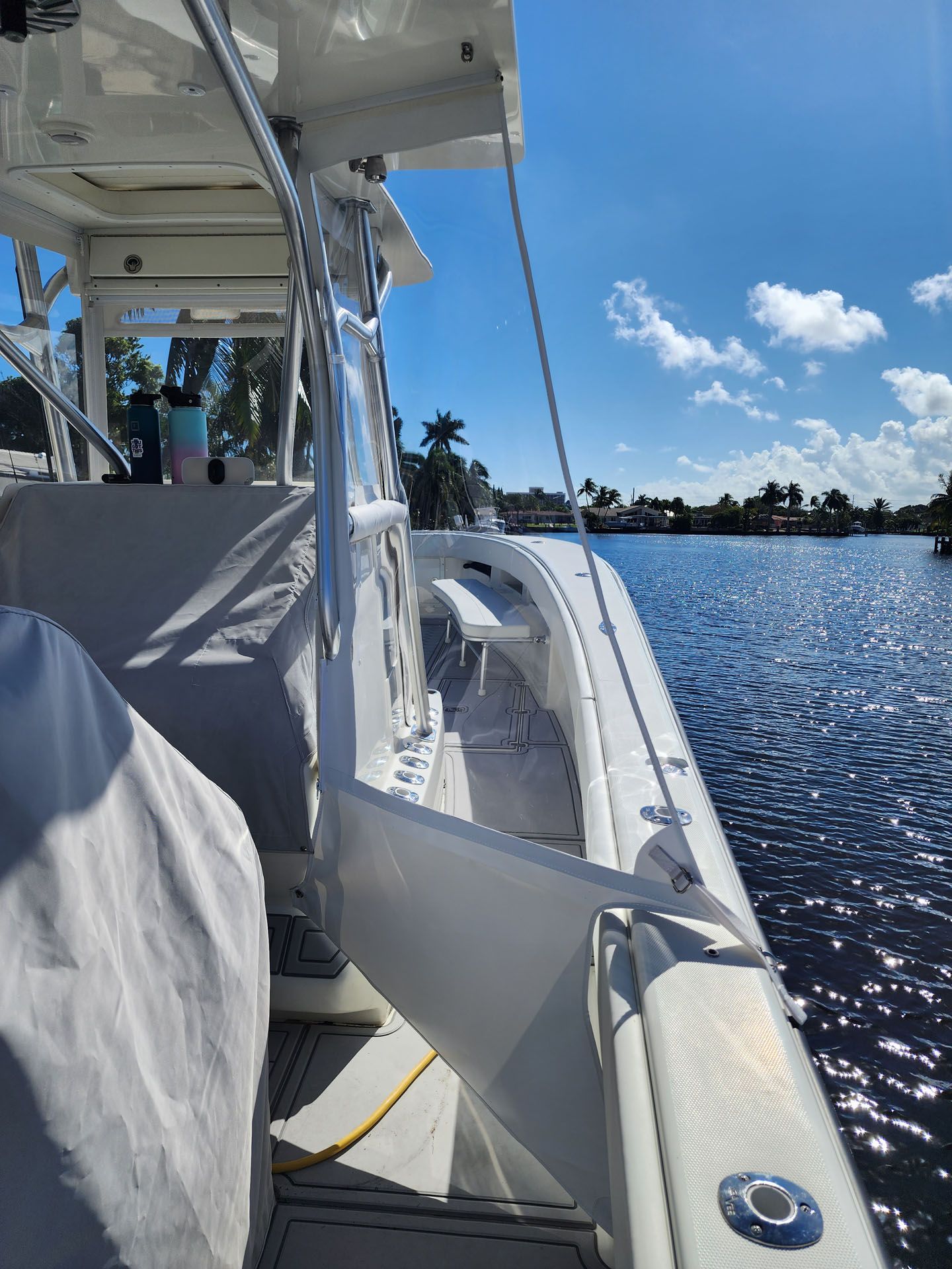 White boat on water, sunny day. Boat's side railing visible, blue sky with some clouds, palm trees in background.