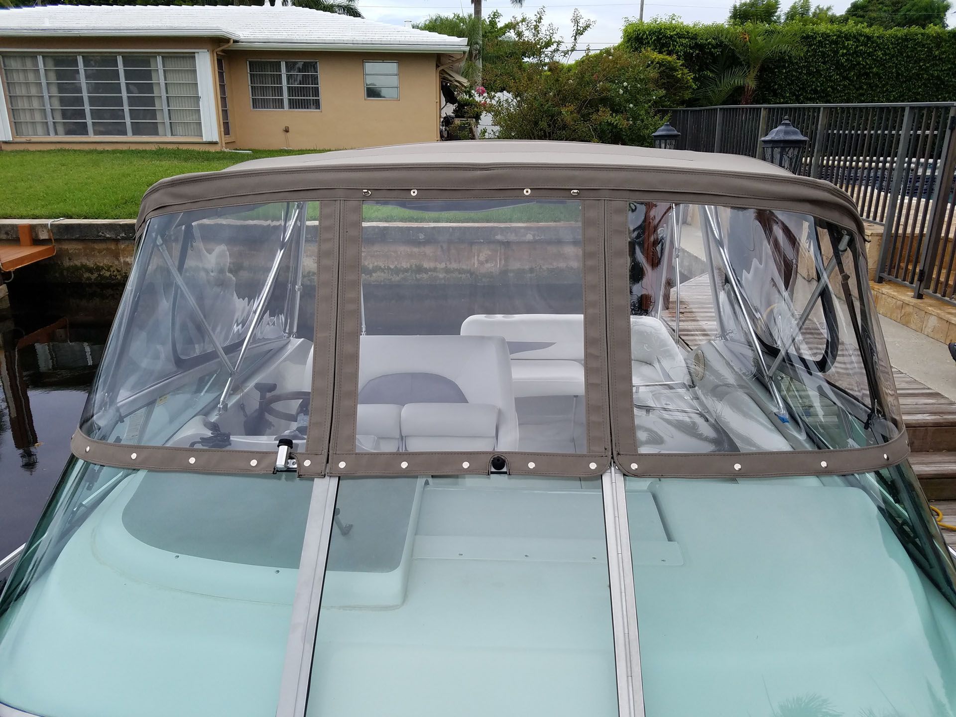 Boat with clear windows and brown fabric covering, docked near a house.