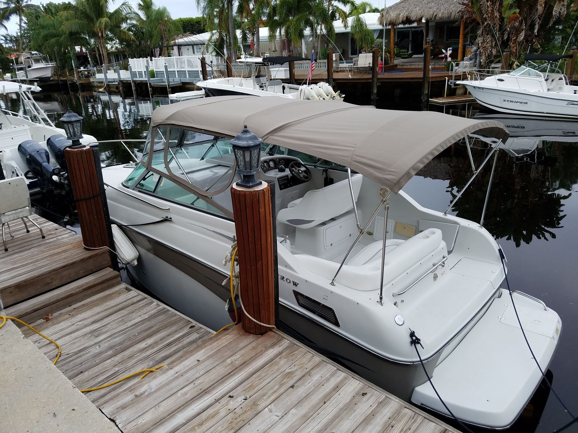 White boat docked at a wooden pier in a canal, beige canopy, two engines.
