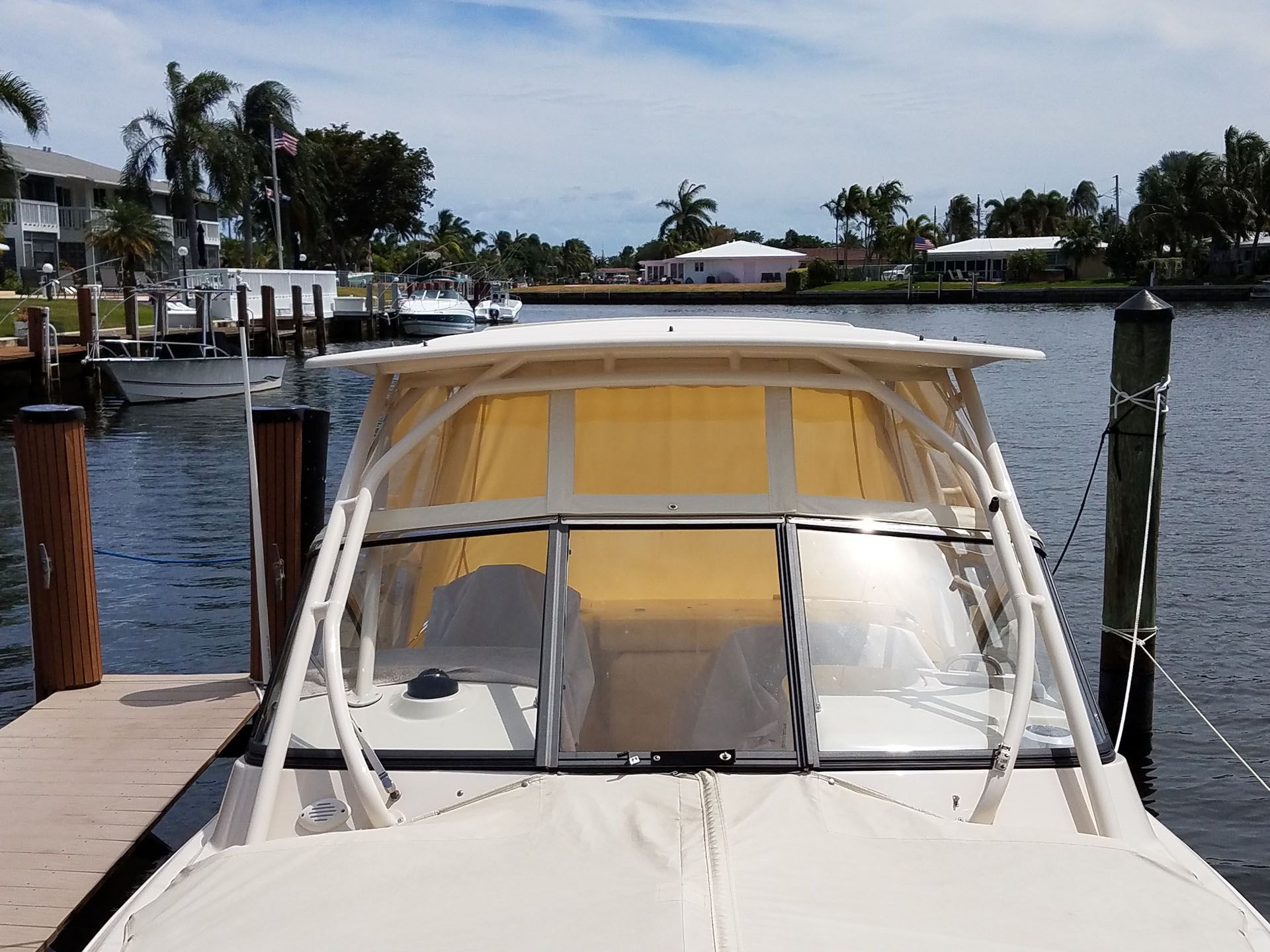 Boat with yellow-tinted enclosure docked at a pier, surrounded by buildings and trees under a bright sky.