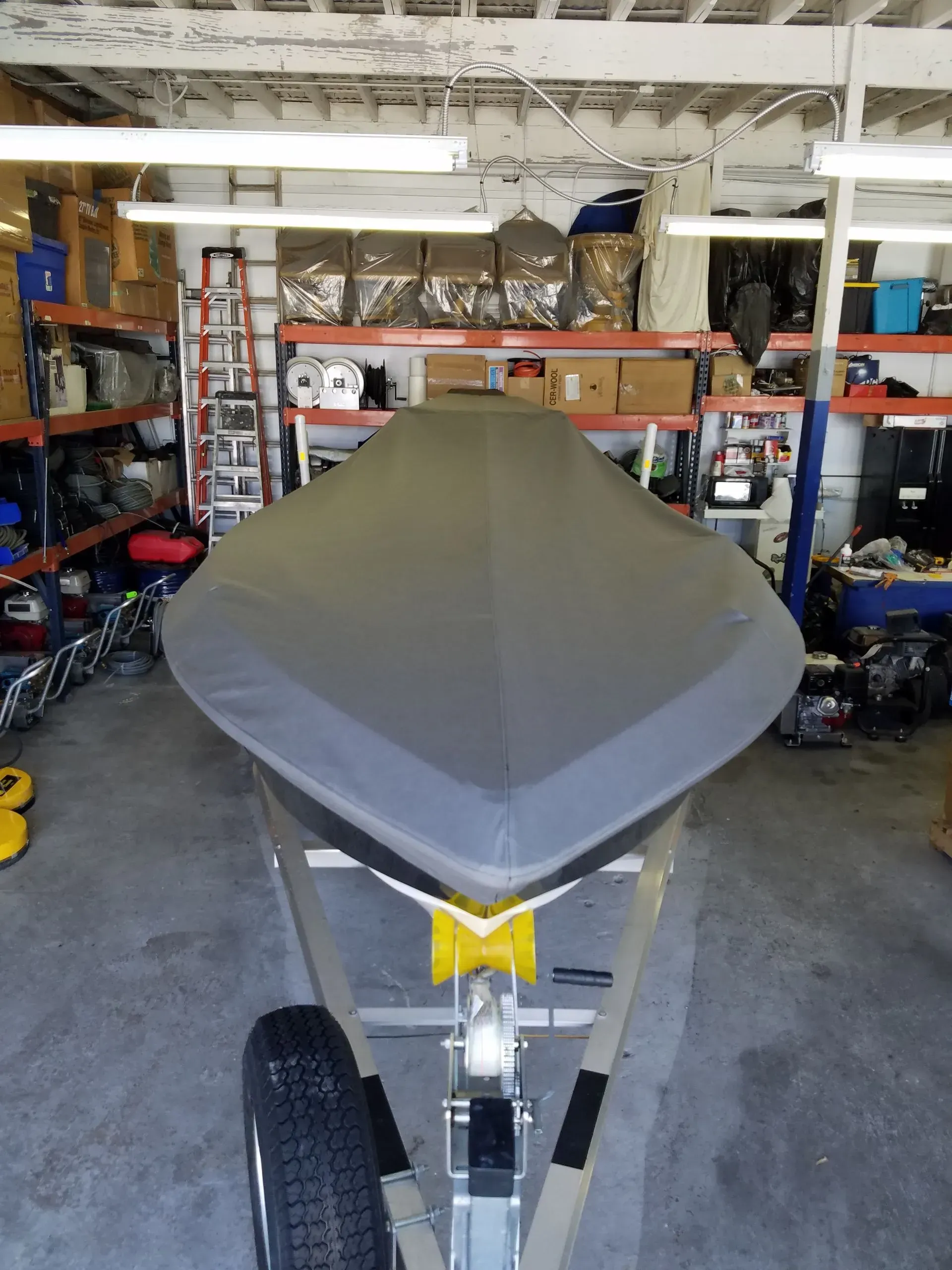 Boat covered with a gray tarp on a trailer, indoors. Yellow trailer arm, storage shelves in background.
