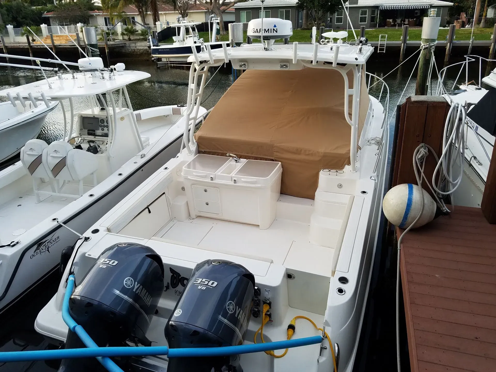White motorboat with dual blue engines docked at a pier. Beige sun cover, white cooler, and life buoy visible.