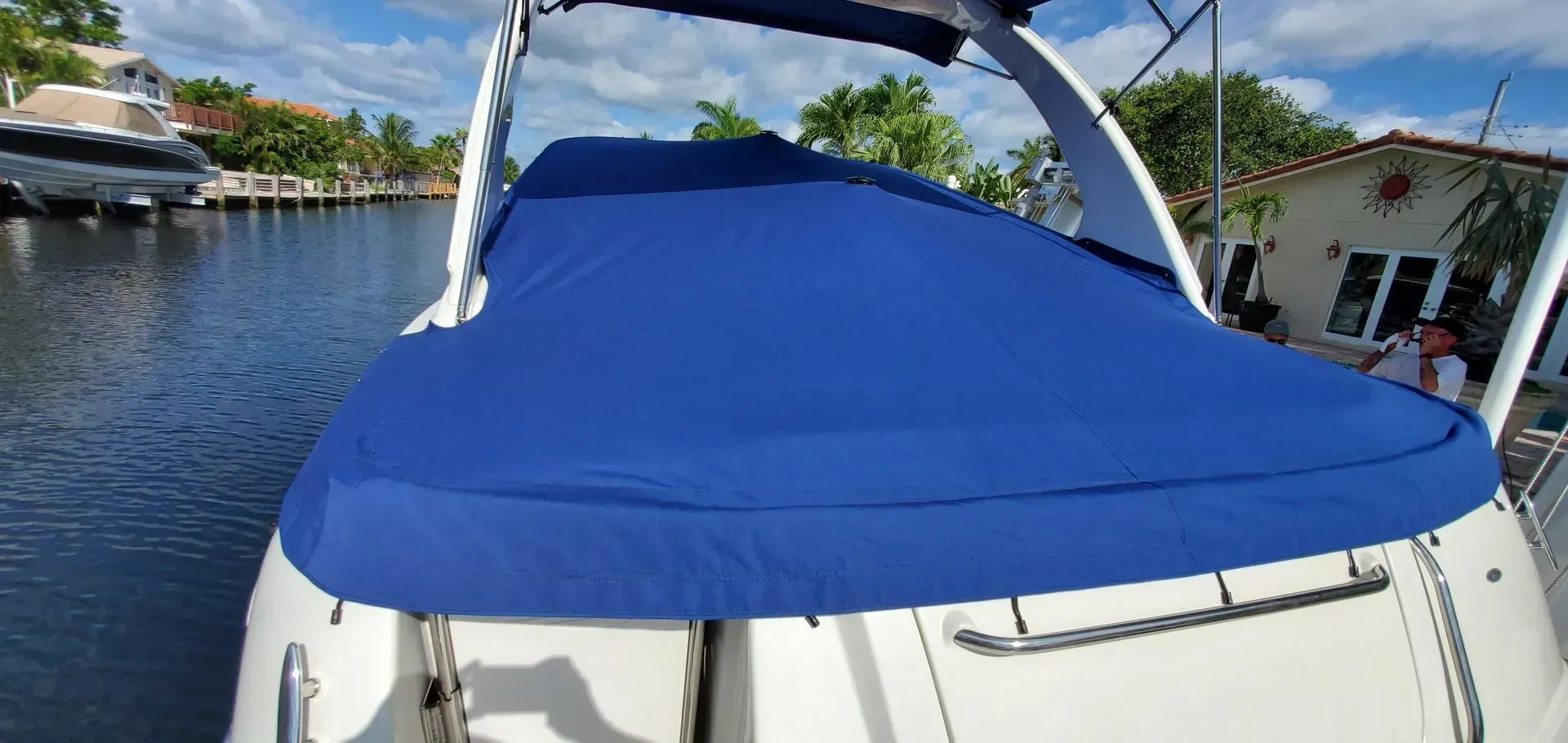 A blue boat cover on a white boat is on a canal with a house in the background.