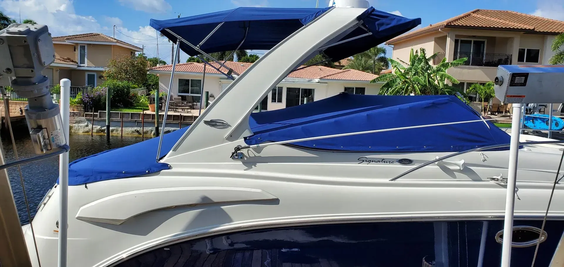 A boat with a blue cover parked by a canal, with houses in the background.