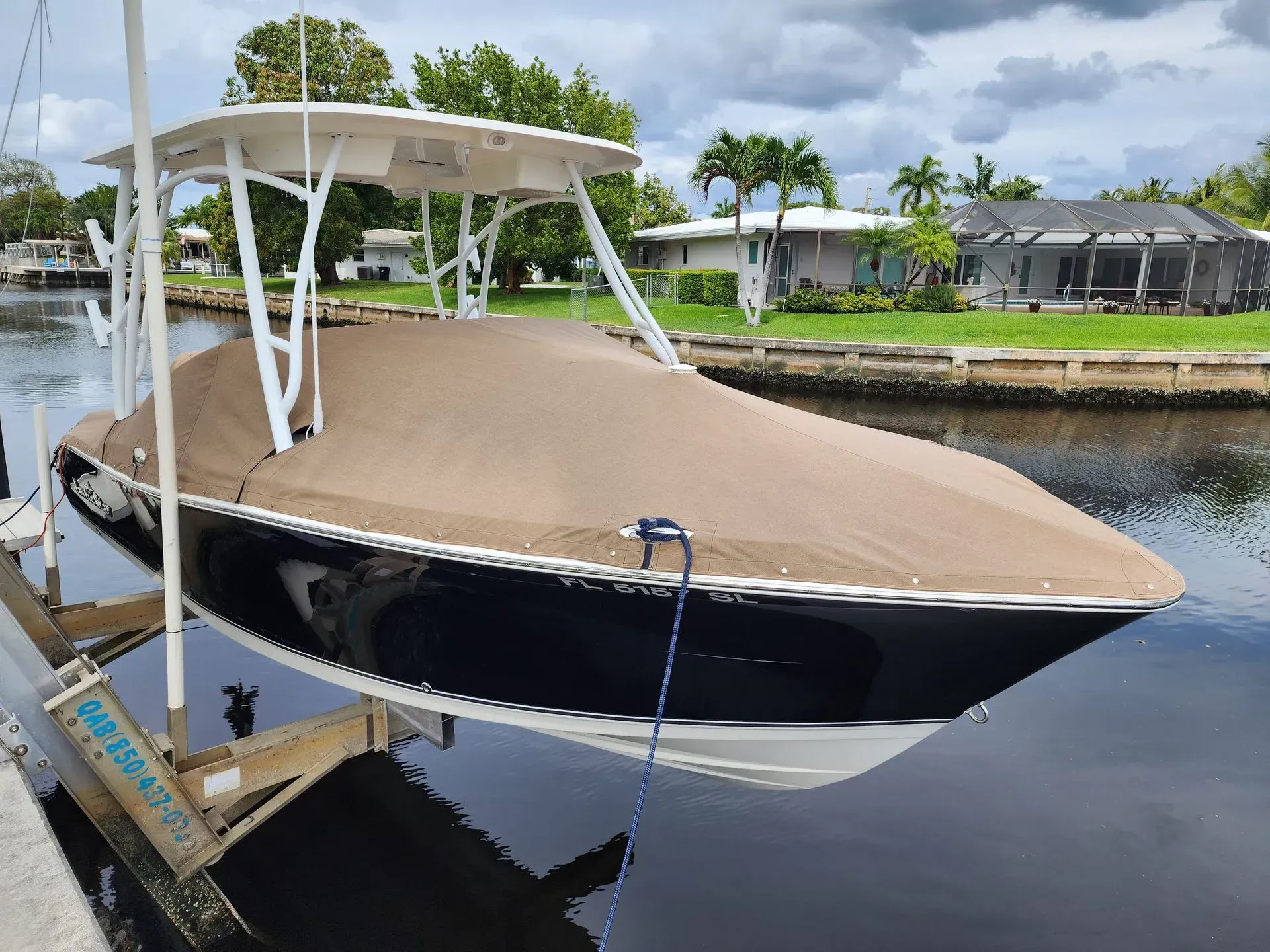 Boat covered in a brown tarp on a lift, in a canal. White T-top, blue hull, green surroundings.