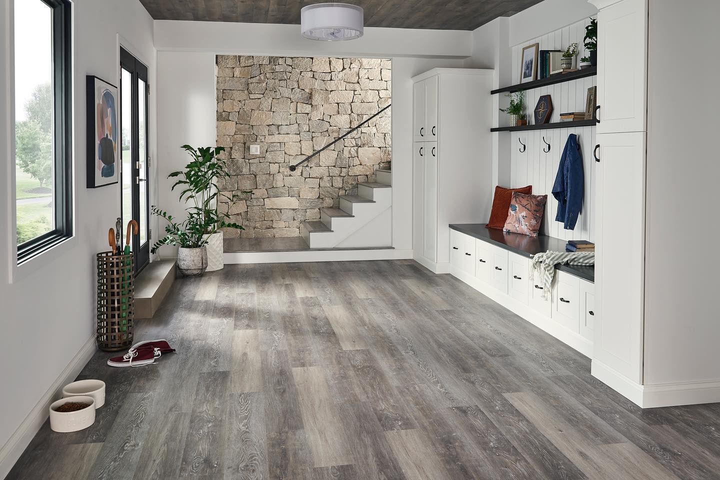 Bright entryway with gray wood floors, stone accent wall, white staircase, and built-in bench with shelves