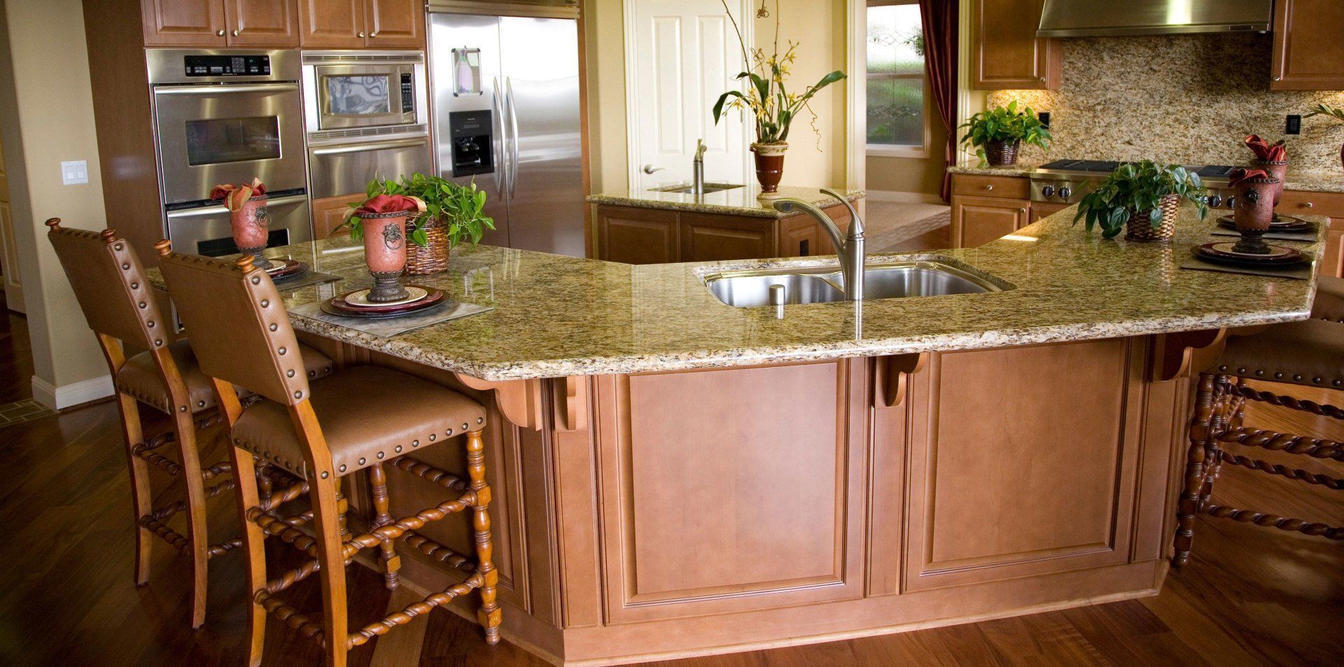 Warm kitchen with granite island, wooden stools, pendant lights, and stainless steel appliances.