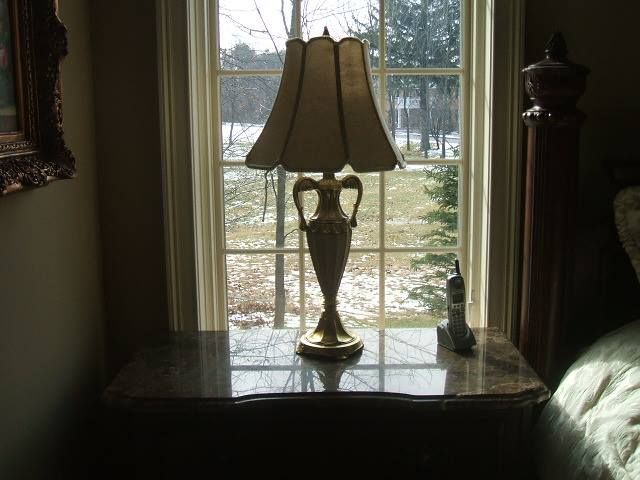 A table lamp on a dark side table by a window overlooking a snowy yard.