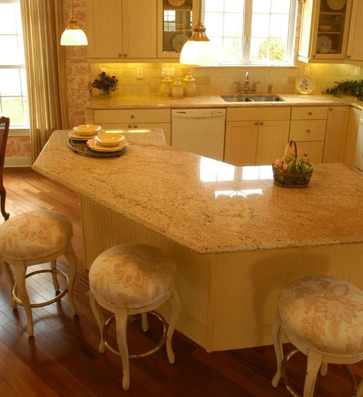 Warm kitchen with granite island, cream cabinets, stools, pendant lights, and flowers by the sink.