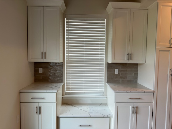 Bright kitchen nook with white cabinets, gray backsplash, and a window with blinds above a countertop.