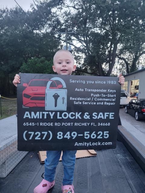 Young child holding an Amity Lock & Safe sign outdoors in Port Richey, Florida. Sign promotes auto keys, repair, and safe services.