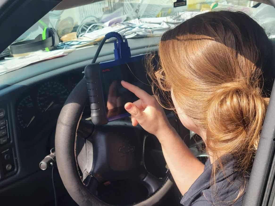 A person with brown hair points at a diagnostic tool plugged into the dashboard of a car.