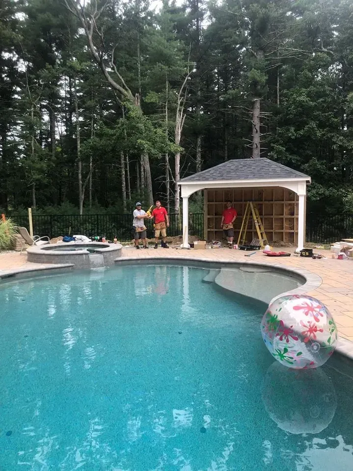 Pool with gazebo under construction; three workers, one holding a drink, with trees in the background.