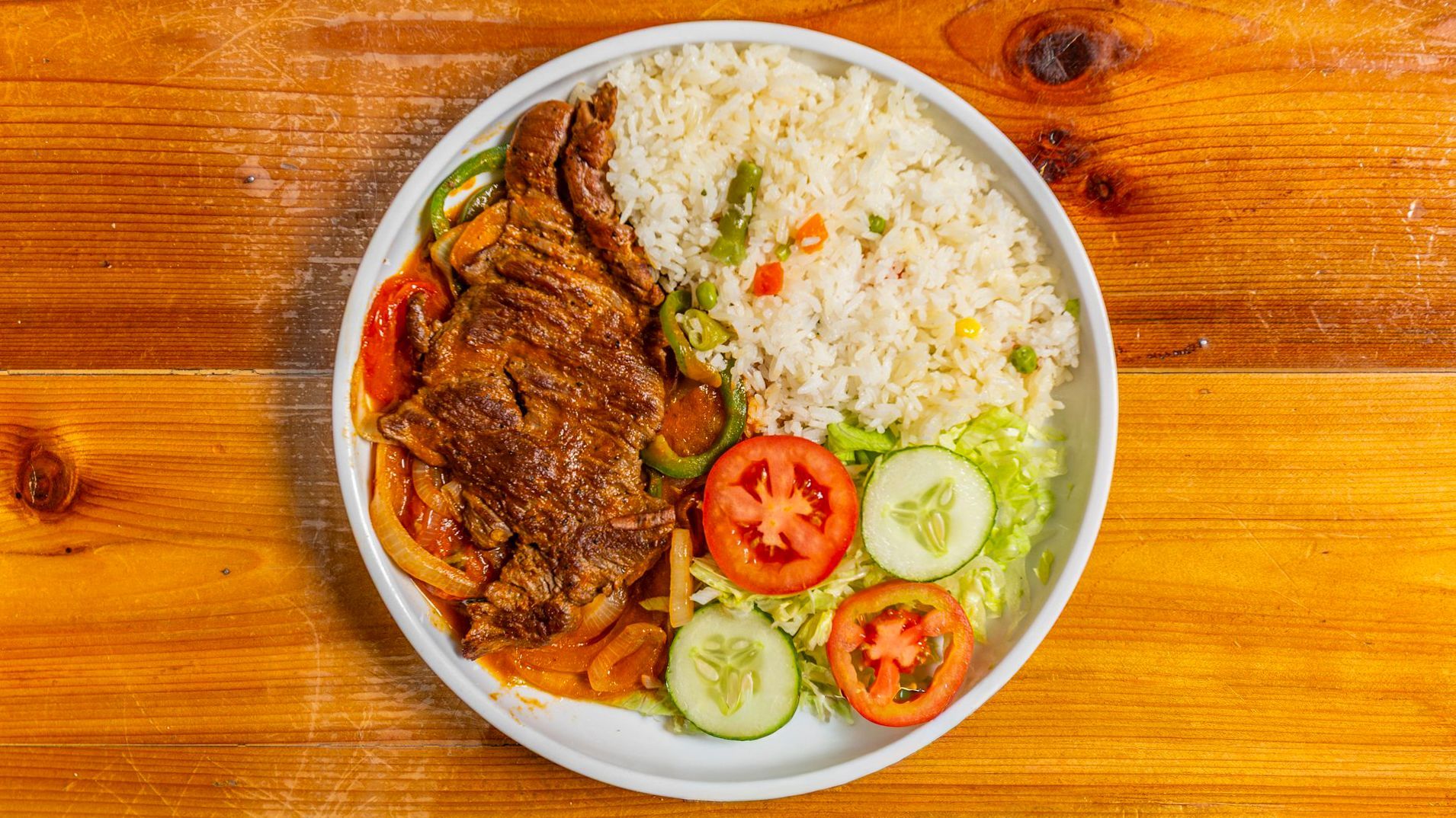 Plate of food: Rice, steak, vegetables (tomato, cucumber, peppers), and lettuce, set on a wooden surface.