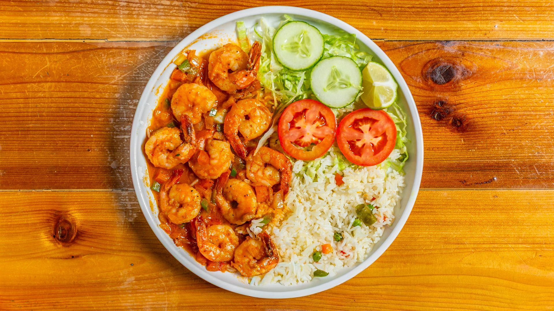 Shrimp dish with rice, tomatoes, cucumber, and lime on a white plate on a wooden table.