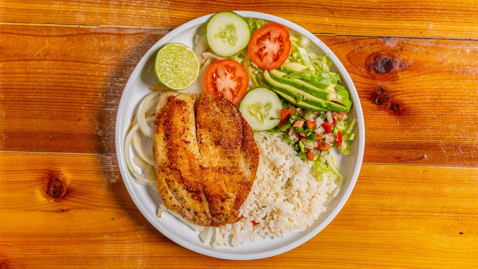 Plate of grilled chicken, rice, salad, avocado, tomato, cucumber, lime, and onion.