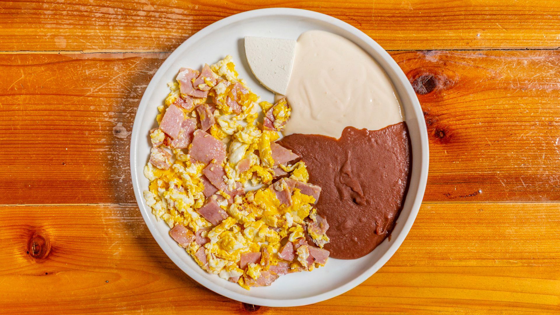 Breakfast plate with scrambled eggs and ham, refried beans, and white cheese.