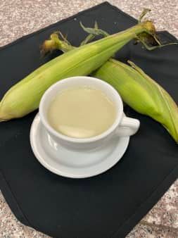 A white cup of creamy corn soup on a saucer, with two ears of corn on a black napkin.