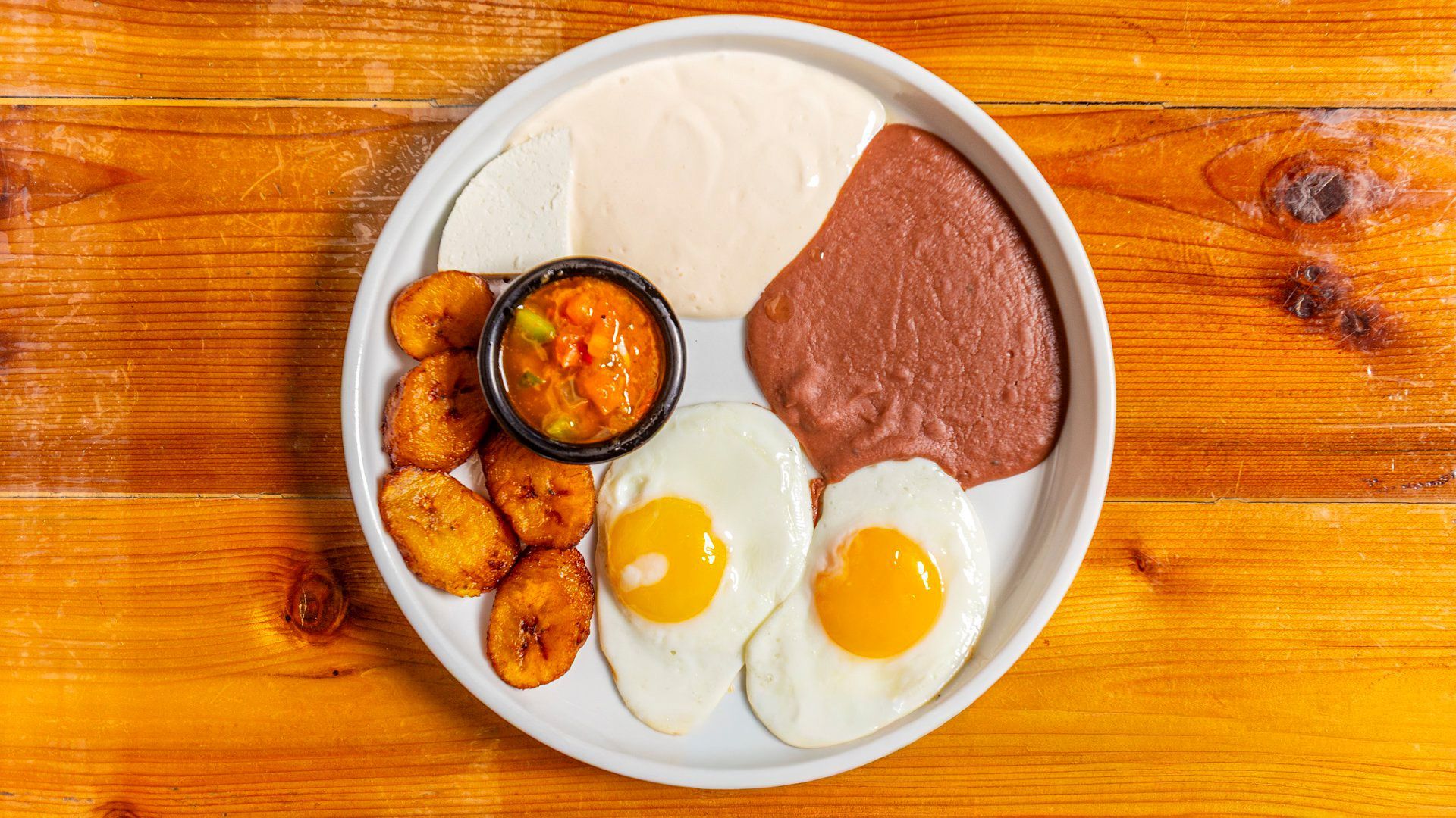 Breakfast plate with fried plantains, eggs, beans, and sauce on a wooden table.