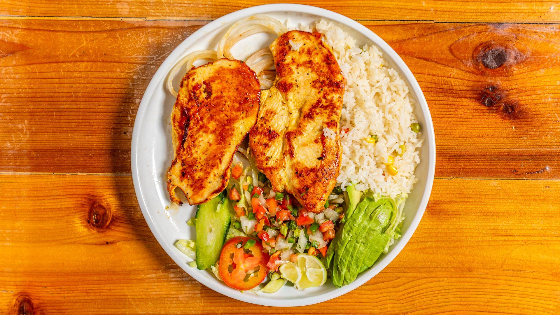 Plate of grilled chicken breasts, rice, avocado, pico de gallo, and onion rings on a wooden table.