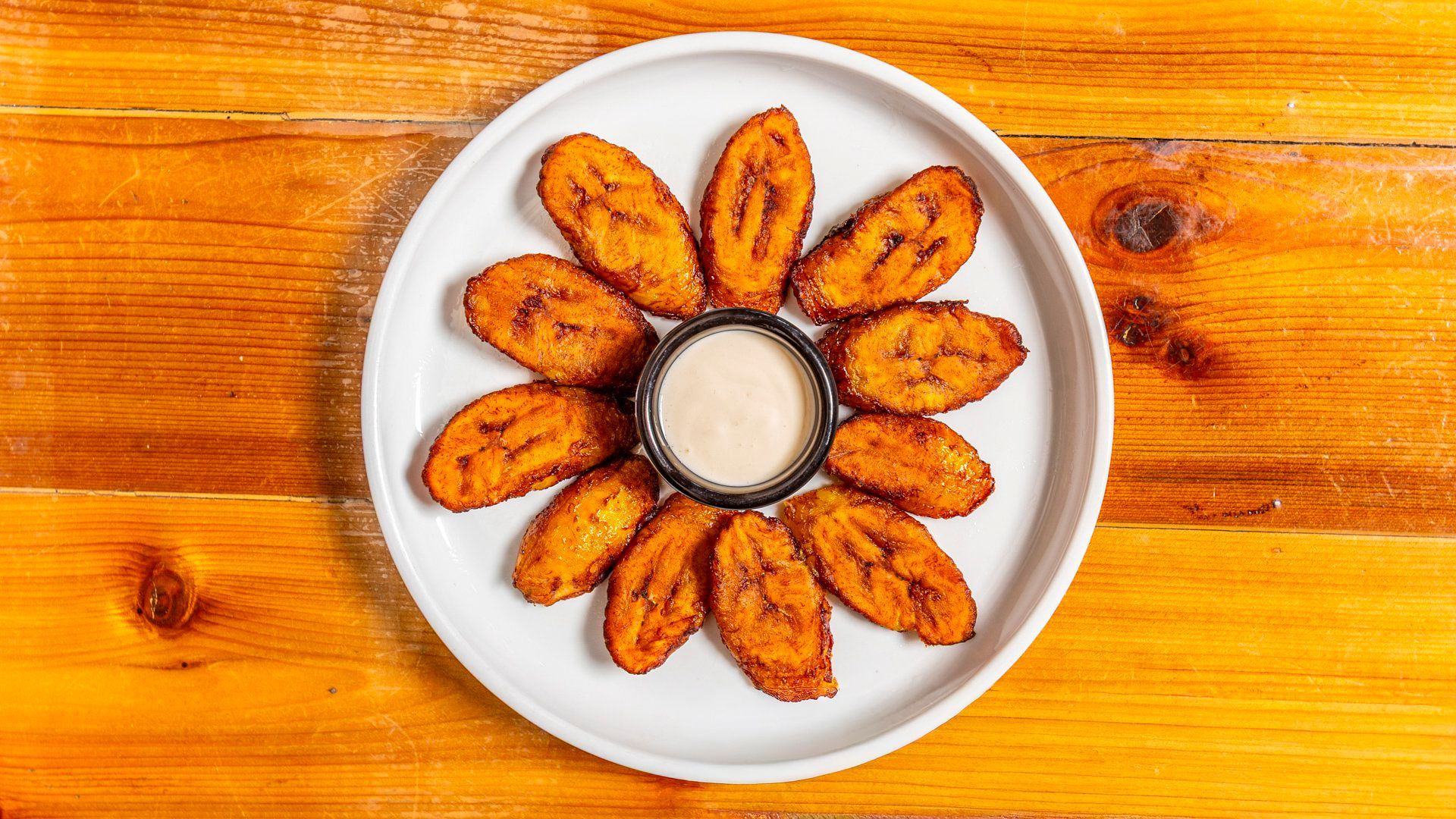 Fried plantain slices arranged in a circle, with a dipping sauce in the center, on a white plate.