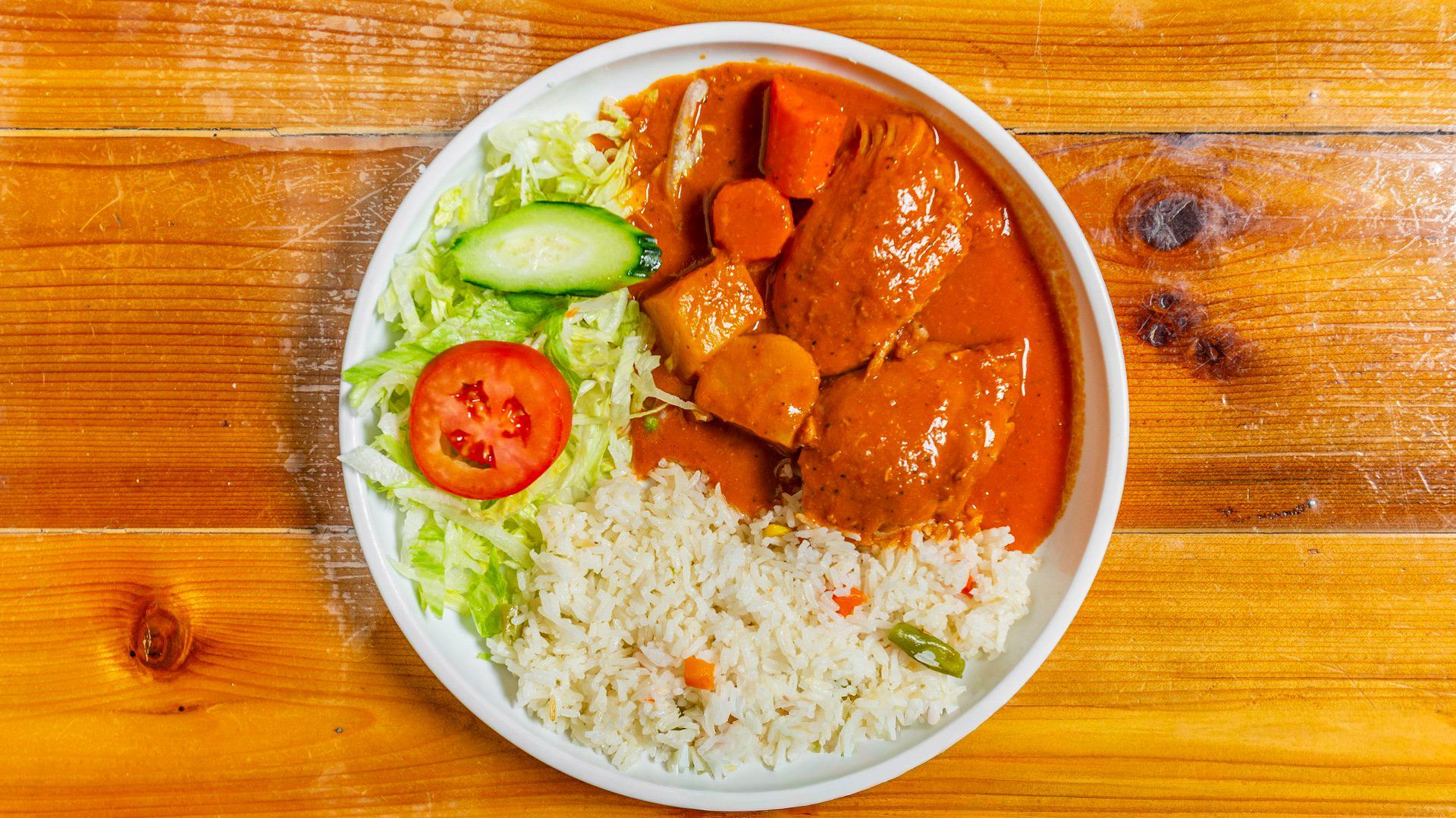 Plate of food: Chicken in sauce, rice, vegetables, on a wooden table.