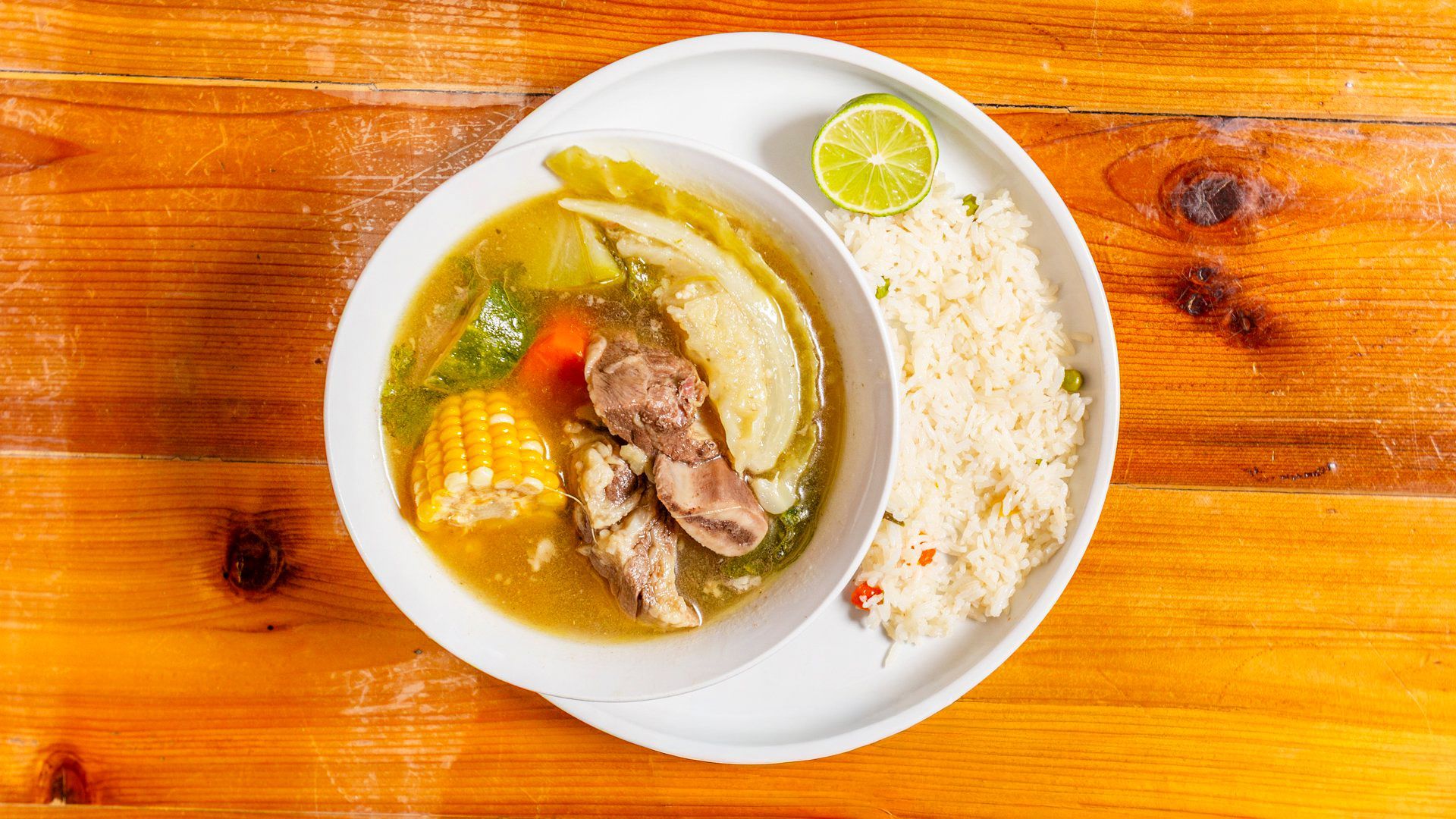 Soup with meat, corn, and greens served with rice and lime on a white plate, wooden table.