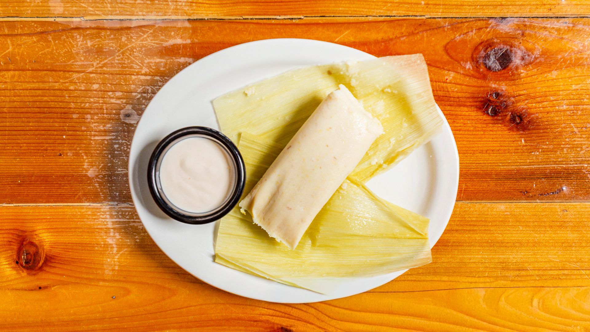 Tamale in corn husk on a white plate, served with a side of cream, on a wood table.