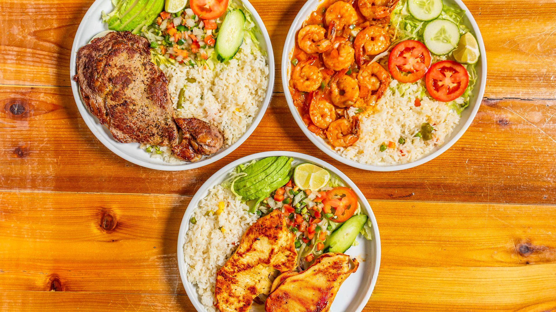 Three plates of food on a wooden surface: steak, shrimp, and fish with rice, vegetables, and avocado.