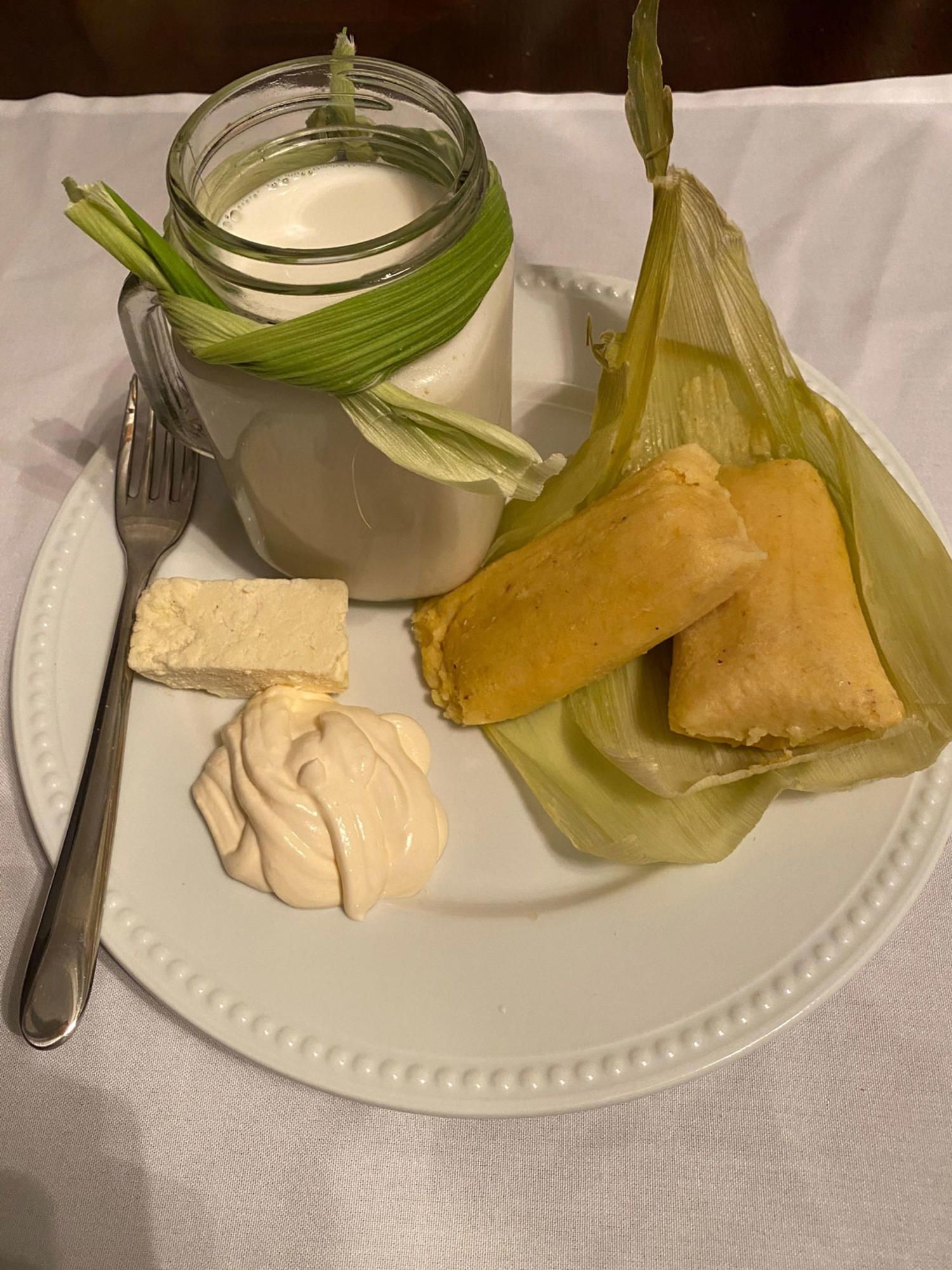 A plate with tamales in husk, drink in a jar, and creamy side dishes.