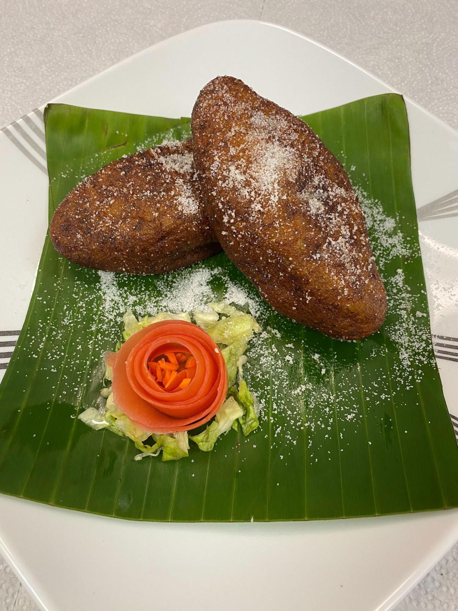 Two fried bread-like food sprinkled with powder on a banana leaf on a white plate, with a tomato rose.