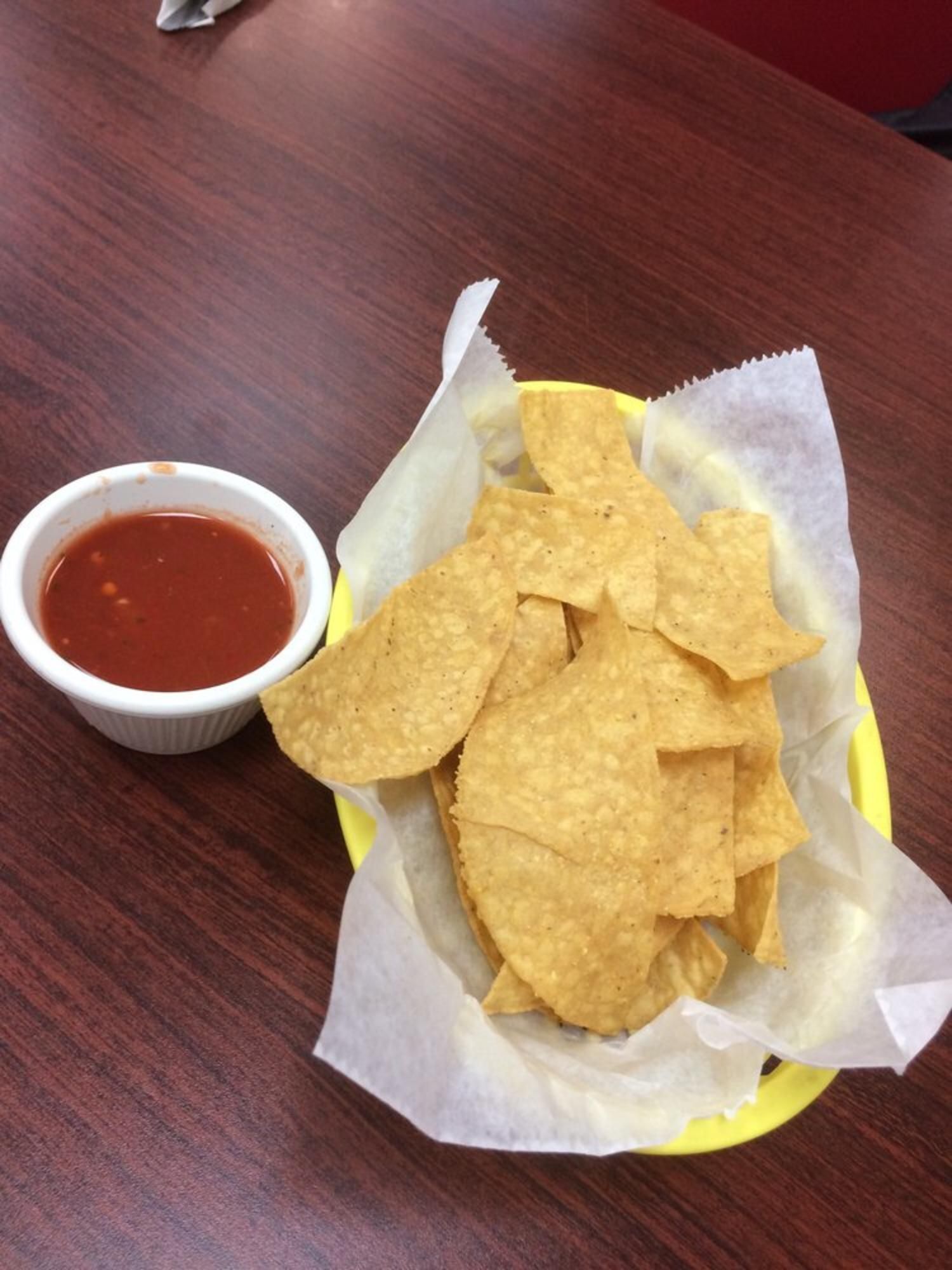 Chips and salsa in a yellow basket on a wooden table. Red salsa in a small white cup.