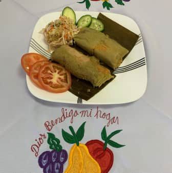 Plate with two tamales, salad, sliced tomato, and cucumber on a table with embroidered fruit and text.