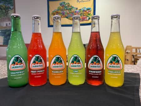 Six bottles of Jarritos soda in various fruit flavors, lined up on a dark surface.