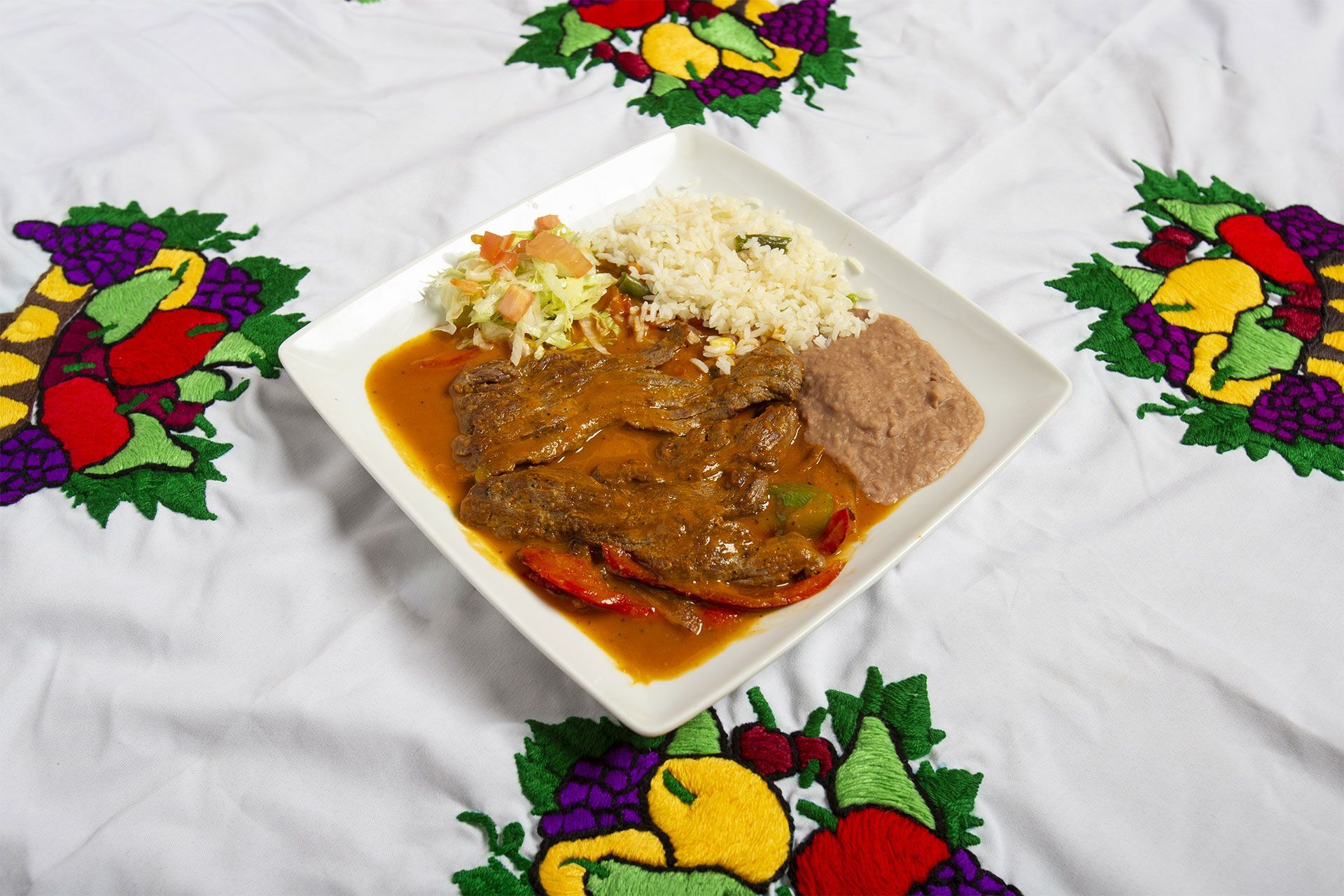 Plate of food on embroidered tablecloth: meat in sauce, rice, refried beans, and salad.