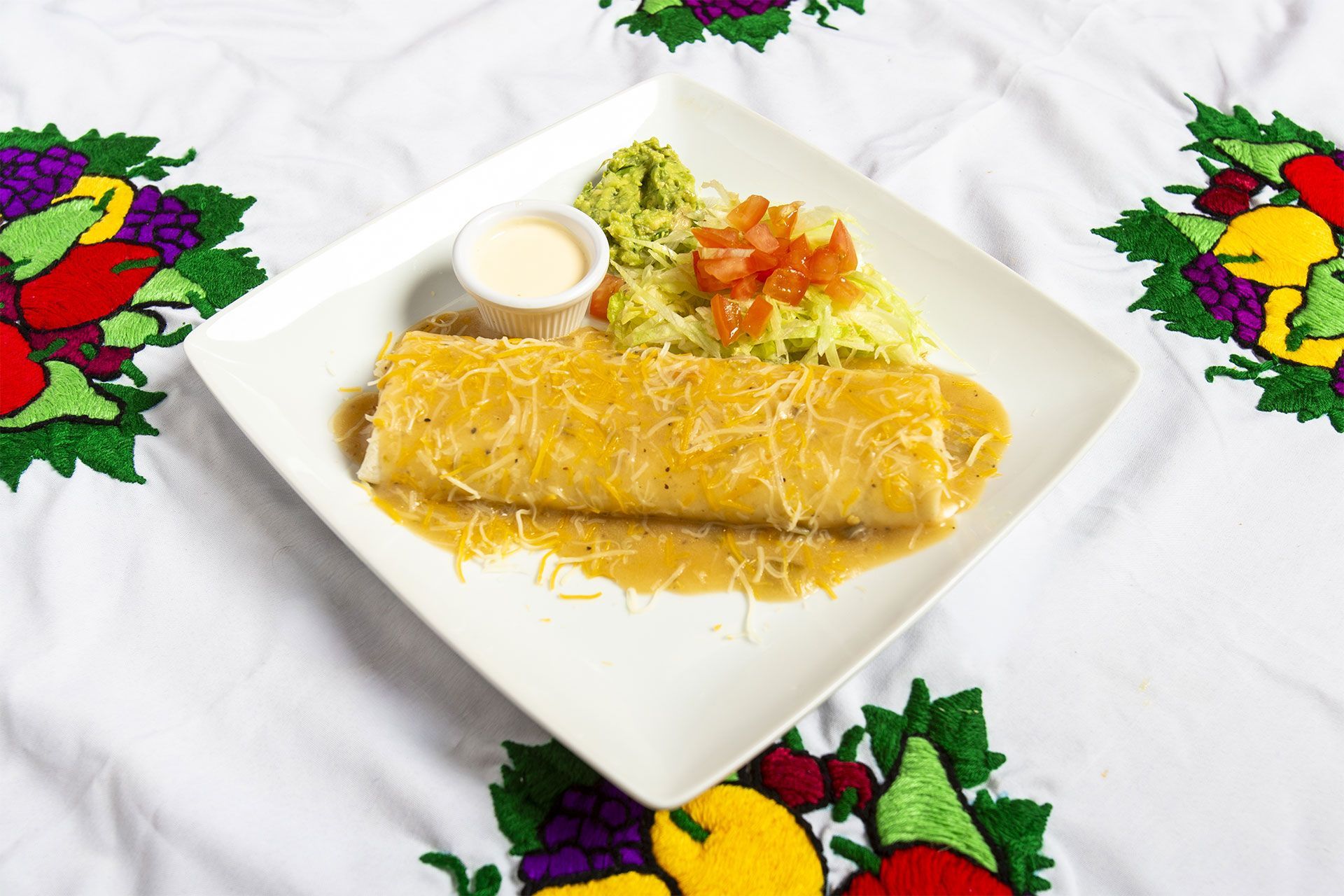 Burrito covered in sauce, served with guacamole, tomato, and crema on a white plate.