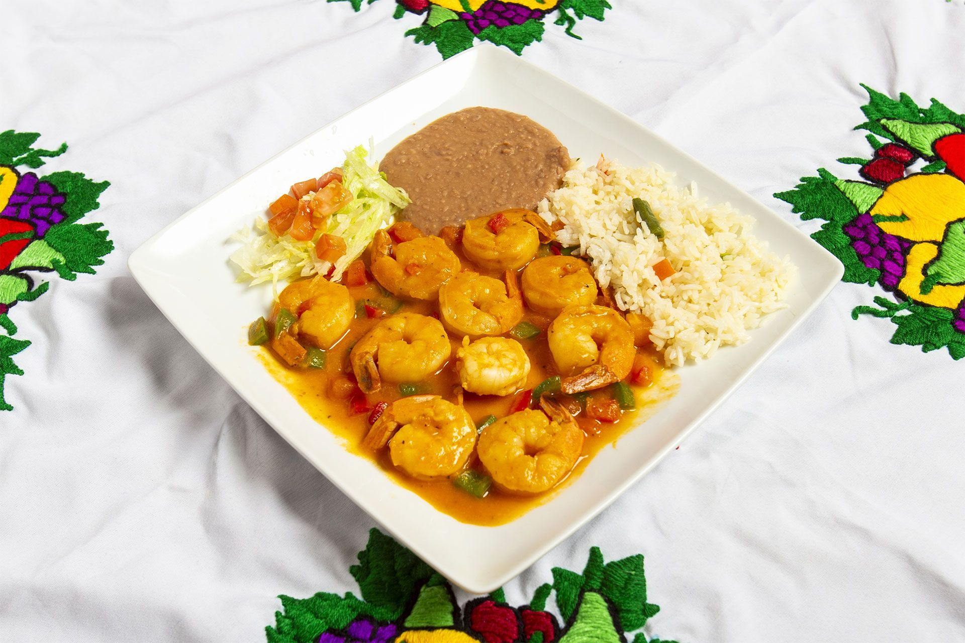 Plate of shrimp with sauce, rice, and beans on a white tablecloth with floral embroidery.
