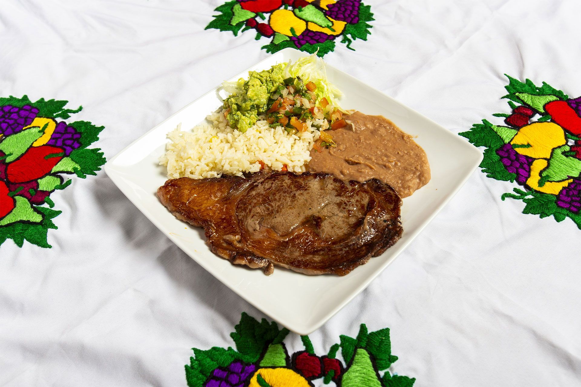 Steak, rice, refried beans, and guacamole on a white plate on a tablecloth with embroidered fruit.