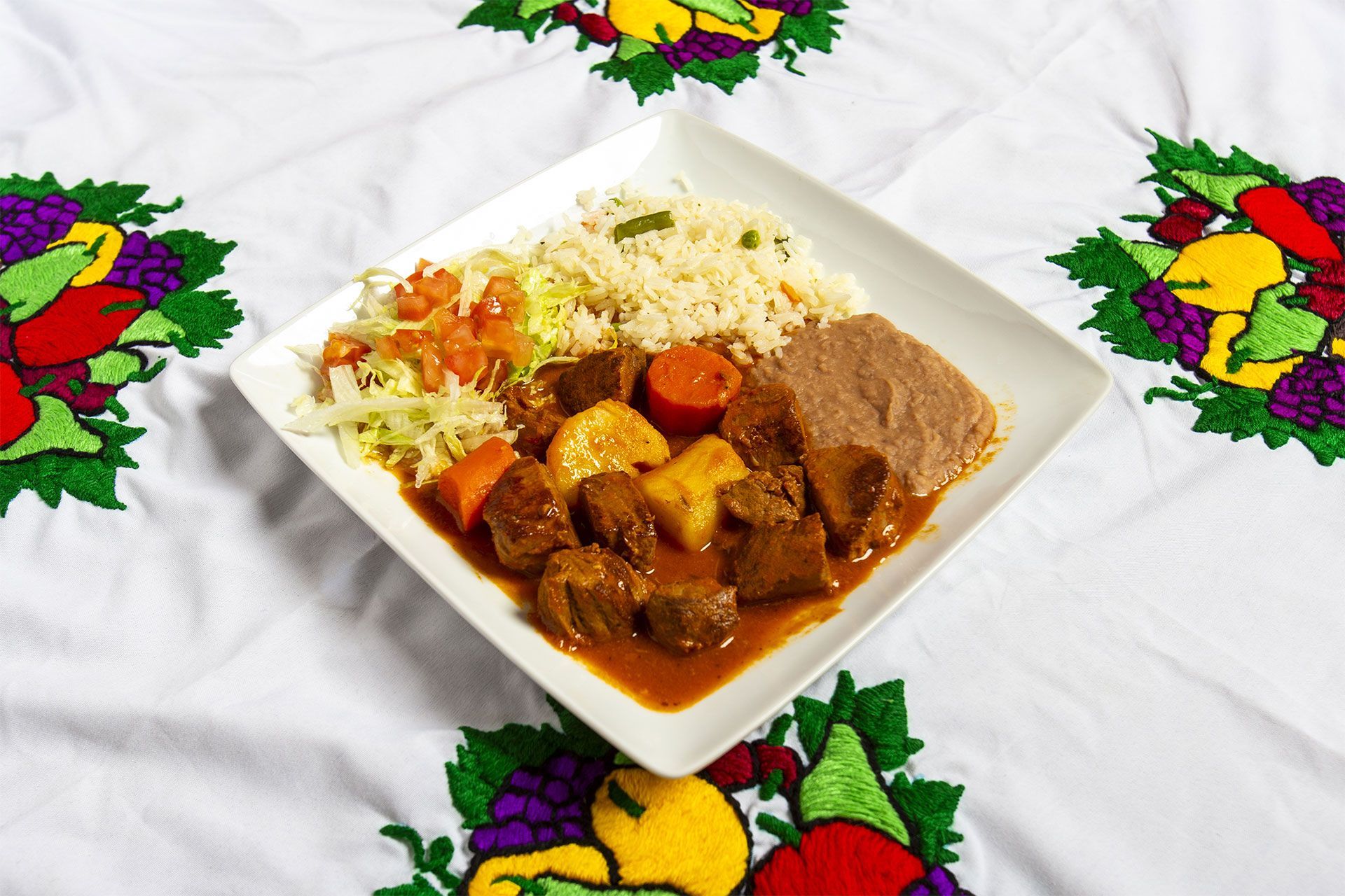 Stew, rice, and beans on a white square plate on a white tablecloth with fruit embroidery.