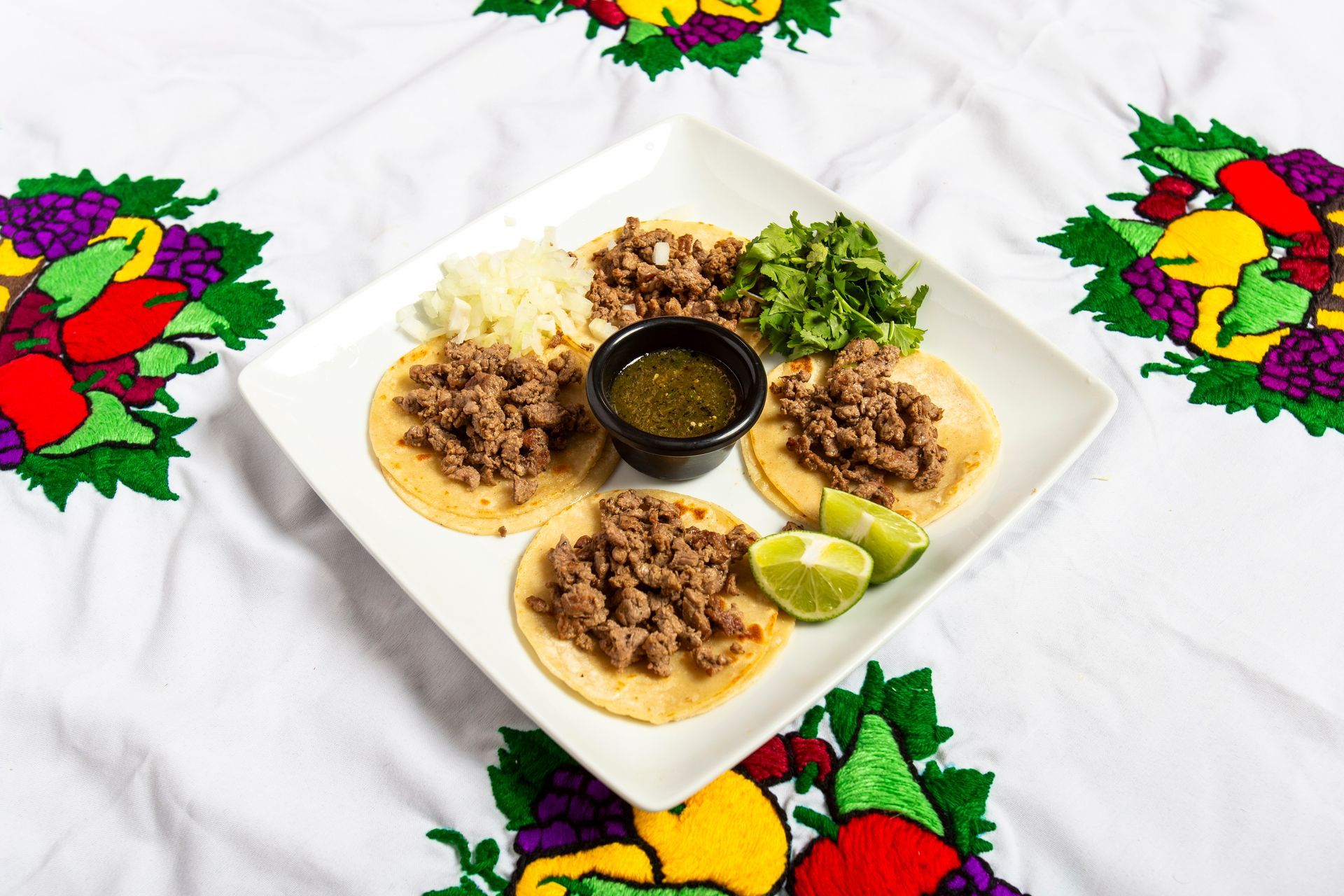 Tacos on a square plate with salsa, cilantro, lime on embroidered tablecloth.
